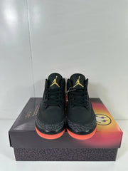 Pair of black Jordan Air Jordan 3 sneakers shown from the front on a patterned shoebox, with gold tongue logos and coral-red outsoles.