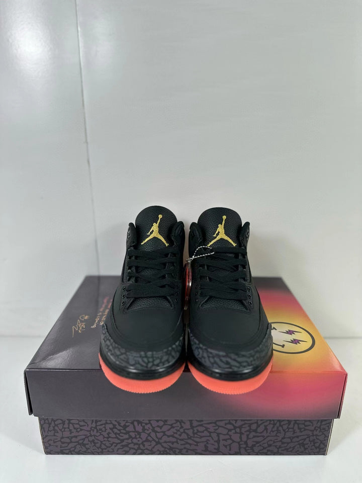 Pair of black Jordan Air Jordan 3 sneakers shown from the front on a patterned shoebox, with gold tongue logos and coral-red outsoles.
