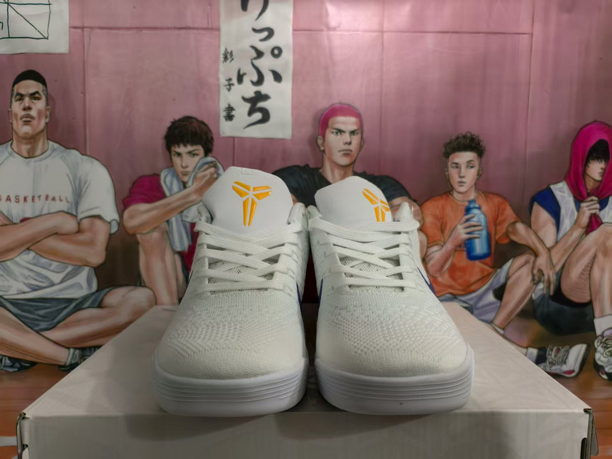 Front view of white basketball sneakers with yellow tongue logos and purple side accents, displayed on a shoebox.