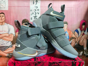 Angled view of grey Nike high-top basketball shoes with strap closures and white-and-gold accents.