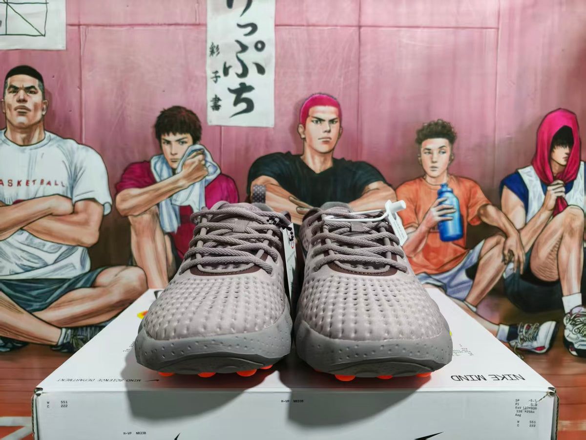 Front view of gray-purple Nike Mind sneakers with orange outsole accents displayed on a Nike shoe box.