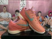 Pair of coral-orange Nike basketball sneakers with green Swoosh branding, pink collars, textured uppers, and cream midsoles displayed on a box.