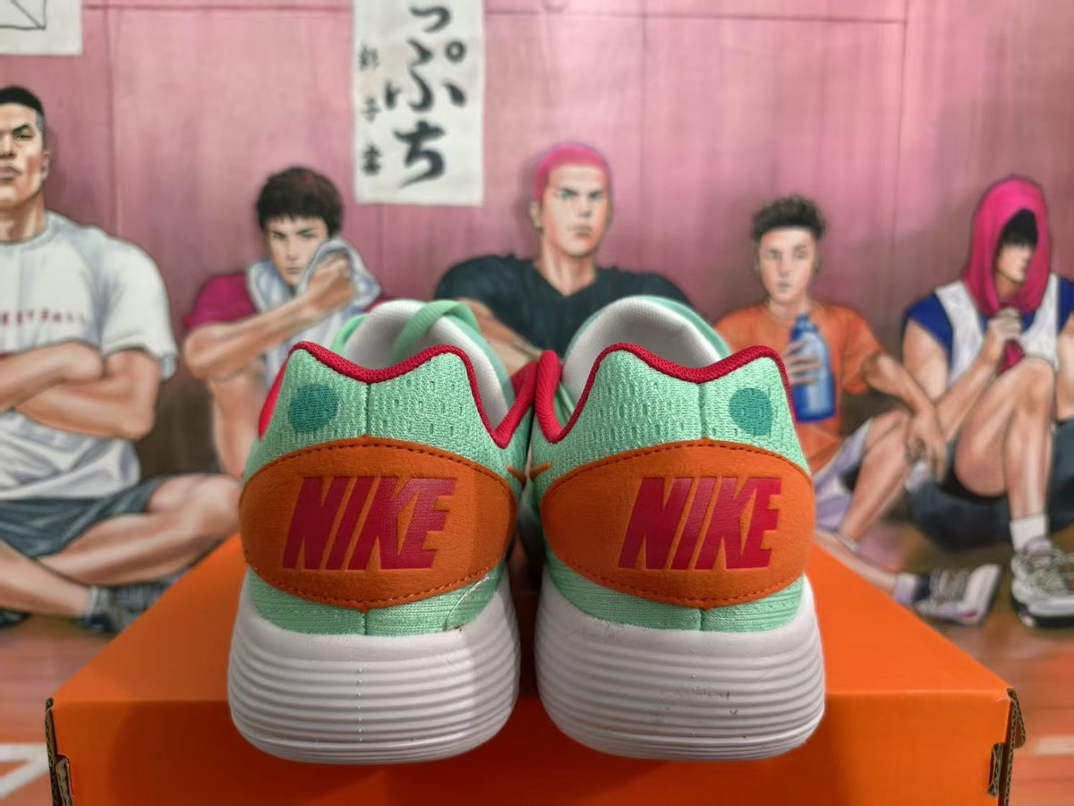 Rear view of mint green Nike sneakers with orange heel panels, red branding, and white textured soles on an orange shoebox.