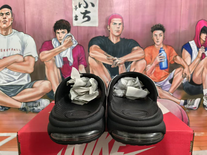 Pair of black Nike slides viewed from the front, showing the cushioned sole and Nike-branded box.