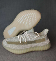 Side and outsole view of silver-toned Yeezy 350 V2 sneakers with an olive stripe and translucent cream sole.
