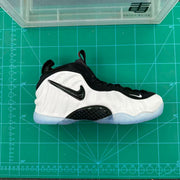 Side view of a white and black Nike Foamposite-style sneaker on a green measuring mat, showing the sculpted upper, black swoosh, and translucent outsole.