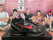 Side view of a Nike basketball sneaker with a red Swoosh, black and olive camouflage upper, and textured midsole.