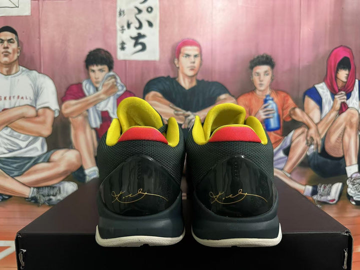 Rear view of dark green and black basketball sneakers with yellow and red collar accents, displayed on a black shoebox.