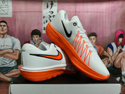 Angled view of white Nike basketball shoes showing black-and-orange Swoosh details and orange soles.