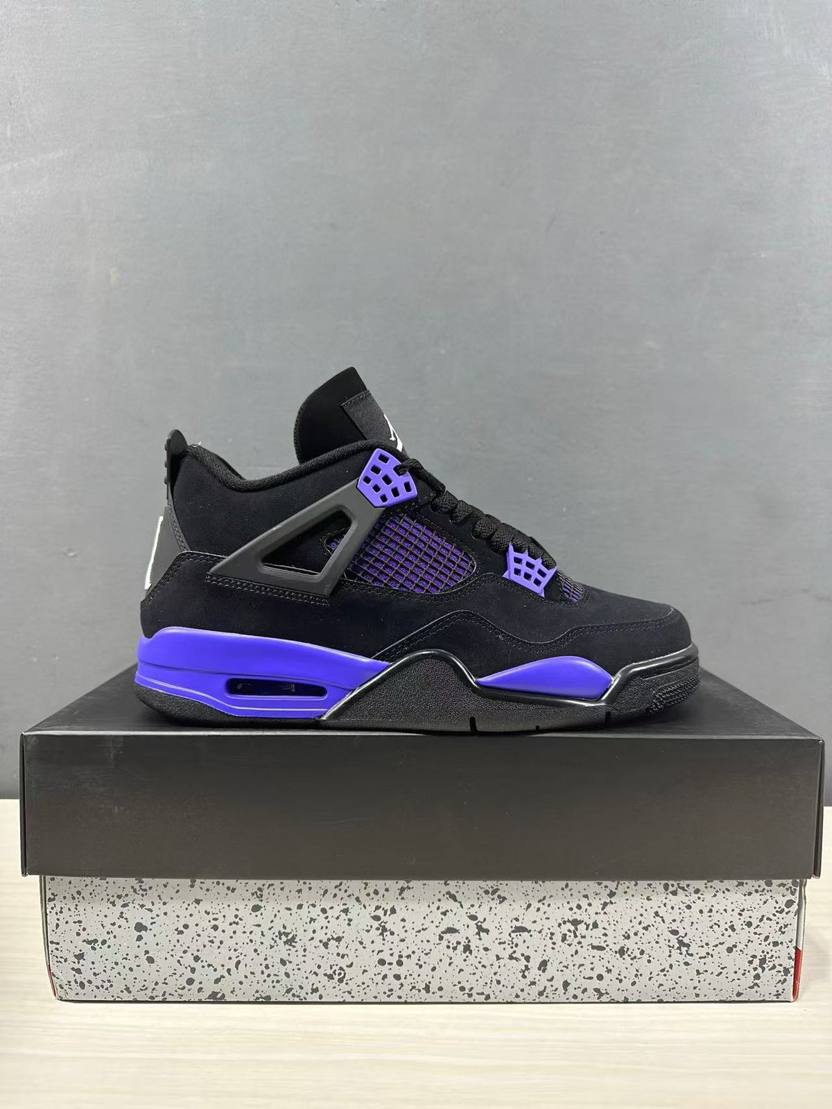Side profile of a black Jordan Air Jordan 4 sneaker with purple-blue accents displayed on a shoebox.
