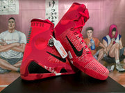 Pair of red high-top basketball sneakers shown from a three-quarter angle, with black Swooshes, textured uppers, and green heel details.