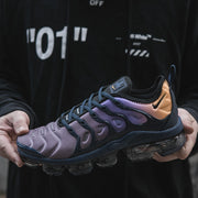 Nike Air VaporMax Plus sneaker held in hand, showing a gray and purple-toned upper, navy overlays, orange heel accent, and segmented sole.
