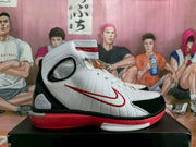 Side profile of a high-top sneaker showing the white upper, black toe and heel panels, red side logo, and red outsole.