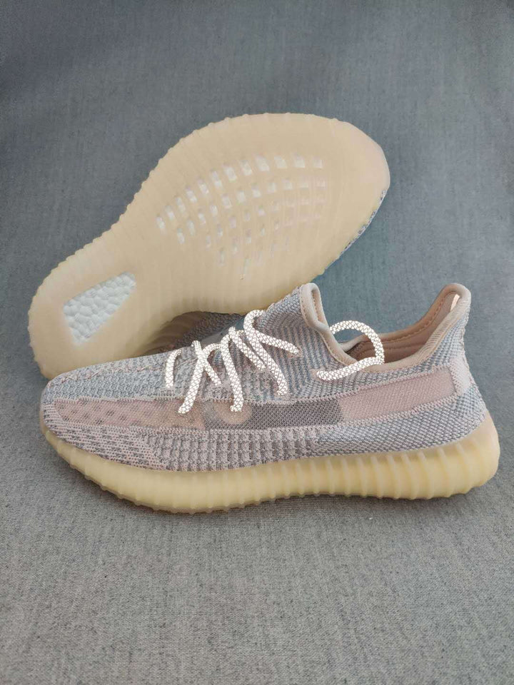 Yeezy 350 V2 style sneaker shown from the side with a grey and pink-toned knit upper, white laces, and a translucent gum-colored sole; outsole visible behind the shoe.