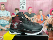 Side view of black and red Under Armour basketball sneakers showing the textured upper, high collar, white outsole sections, and speckled midsole detail.