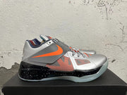 Side view of a silver Nike KD basketball shoe with an orange swoosh, patterned side panel, and speckled black midsole.