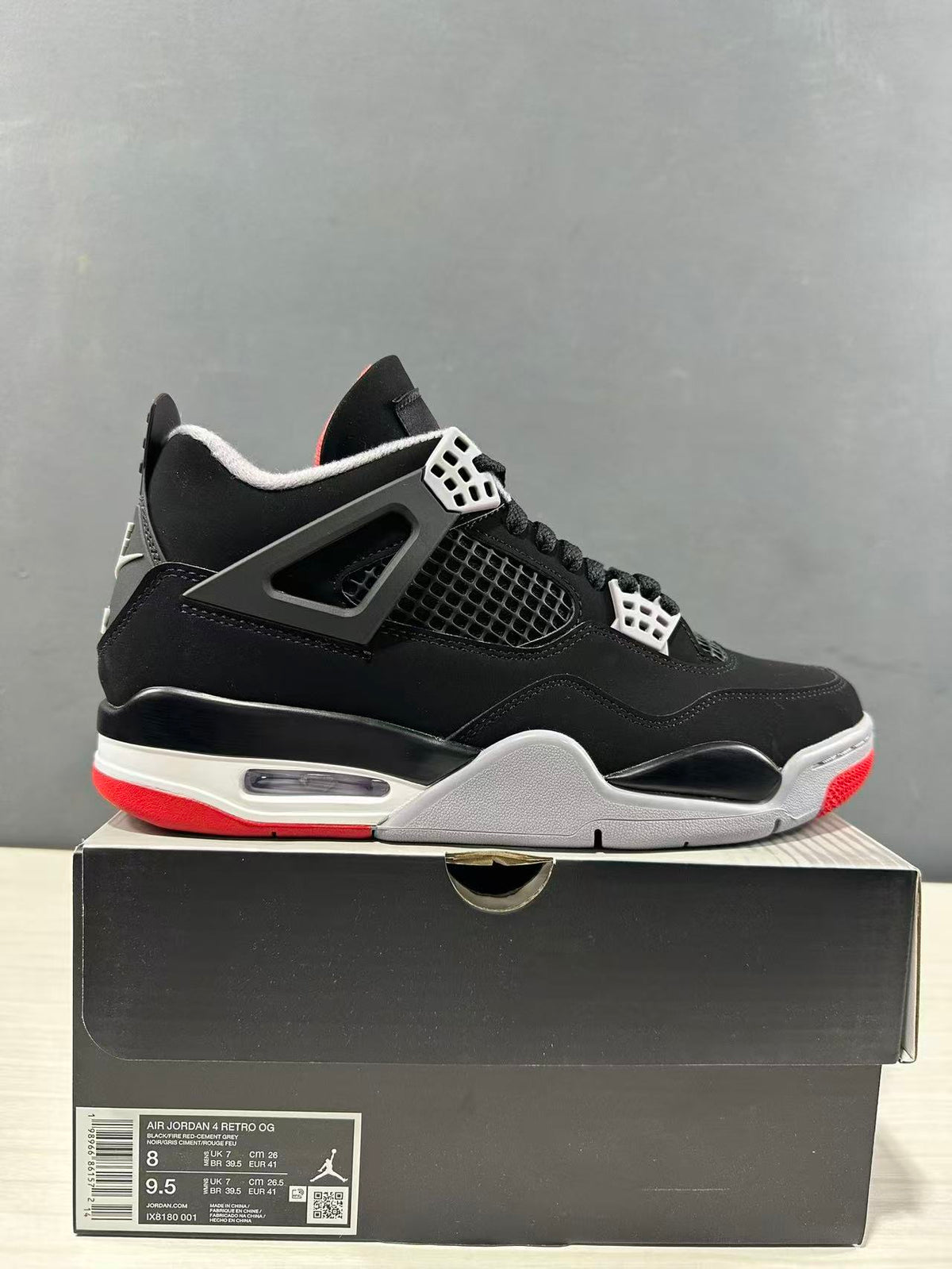 Side view of black and red Jordan Air Jordan 4 sneakers resting on a black shoebox; the box label shows a photographed sample in EU 41 and style code IX8180 001.