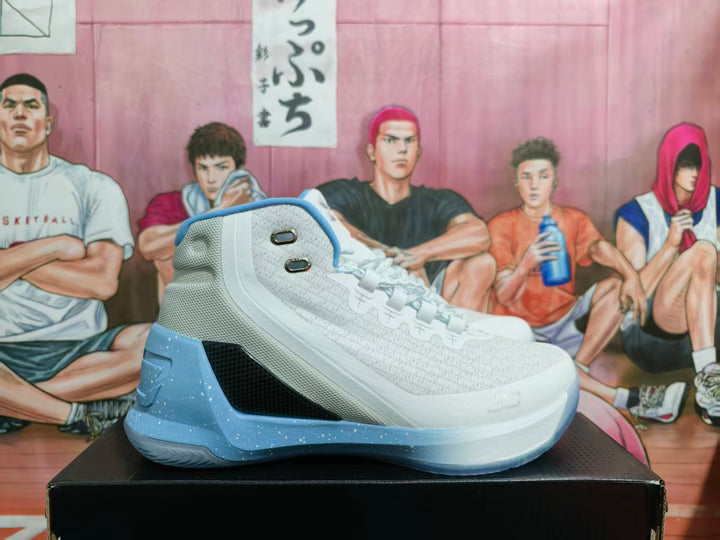 Side view of a white and light blue Under Armour high-top basketball sneaker displayed on a black box.