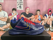 Side view of navy basketball sneakers with red laces, yellow side swoosh detailing, and contrasting heel panels on a shoebox.