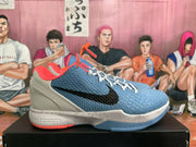 Side view of a blue-grey sneaker with a black Swoosh, orange-red trim, white speckled midsole, and visible Nike Zoom branding.