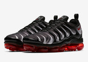 Angled view of a pair of black and gray patterned Nike Air VaporMax Plus sneakers with red soles.