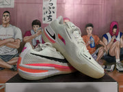 Angled view of two white Nike basketball sneakers displaying black Swoosh branding, pink and coral accents, and translucent outsoles.