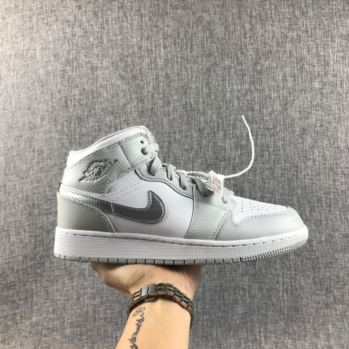 Side view of a white and silver Jordan Air Jordan 1 Mid GS sneaker held against a gray fabric backdrop.
