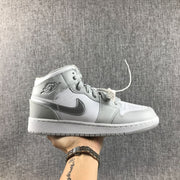 Side view of a white and silver Jordan Air Jordan 1 Mid GS sneaker held against a gray fabric backdrop.