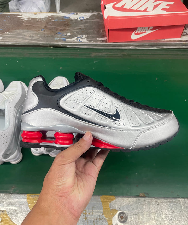 Silver and black Nike Shox-style sneaker with red heel cushioning columns, shown beside a red Nike shoebox.