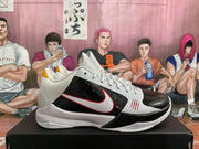 Pair of white and black Nike basketball sneakers displayed on a black shoebox, showing red-edged side branding and yellow heel accents.