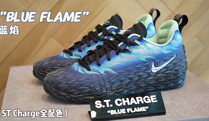 A pair of blue and black Nike S.T. Charge sneakers with black laces, textured dark overlays, visible Swoosh logos, and a Blue Flame display placard.