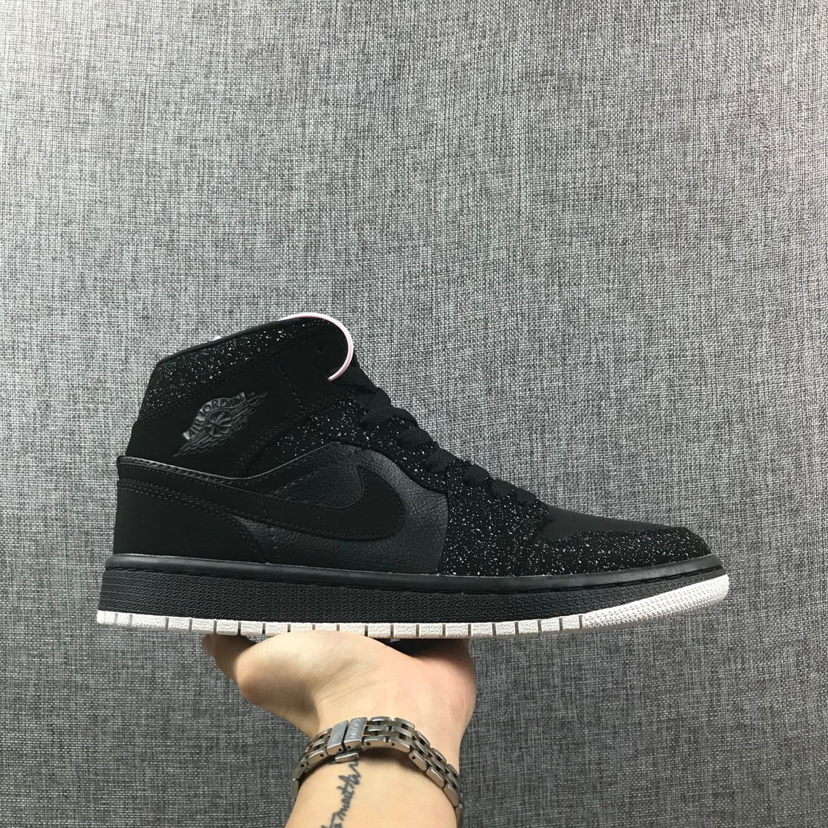 Side profile of a black Air Jordan 1 Mid sneaker with a white outsole and speckled black overlays.