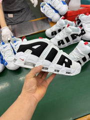 White and black Nike sneakers shown in a workshop setting, with additional white and blue shoes in the background.