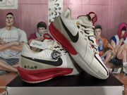 Angled view of a pair of white, black, and red basketball sneakers displaying the side branding, red midsoles, and textured outsole.