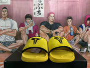 Front view of yellow slide sandals with black upper logos, displayed on a black box.