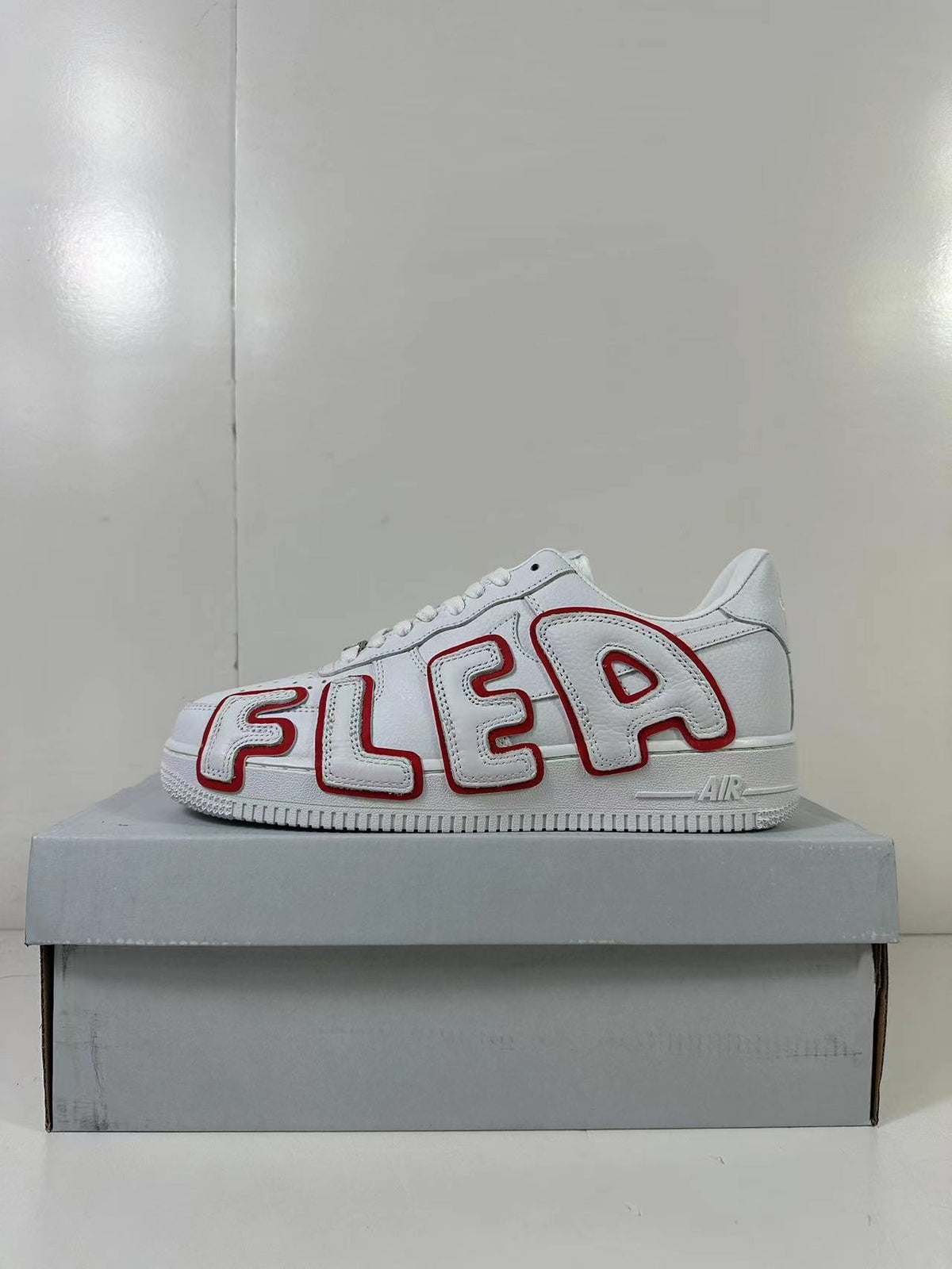 Side view of a white low-top sneaker with oversized red-outlined AIR lettering, displayed on a gray shoebox.