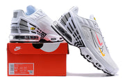 White Nike Air Max Plus sneakers with black accents and pink and yellow graphics, displayed on a red shoe box.