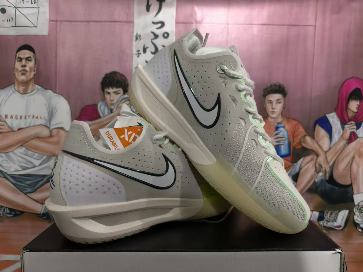 Gray-green Nike basketball sneakers displayed on a shoebox, showing black Swoosh branding, perforated panels, and pale green accents.