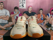 Front view of cream Nike sneakers showing green Swoosh details, orange laces, and orange-tinted soles.