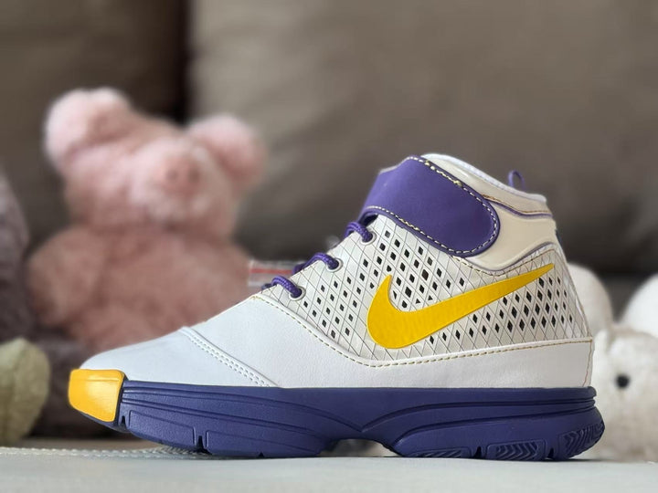 Side profile of a white high-top kids' basketball sneaker with purple ankle overlay, yellow Swoosh, patterned side panel, and dark purple outsole.