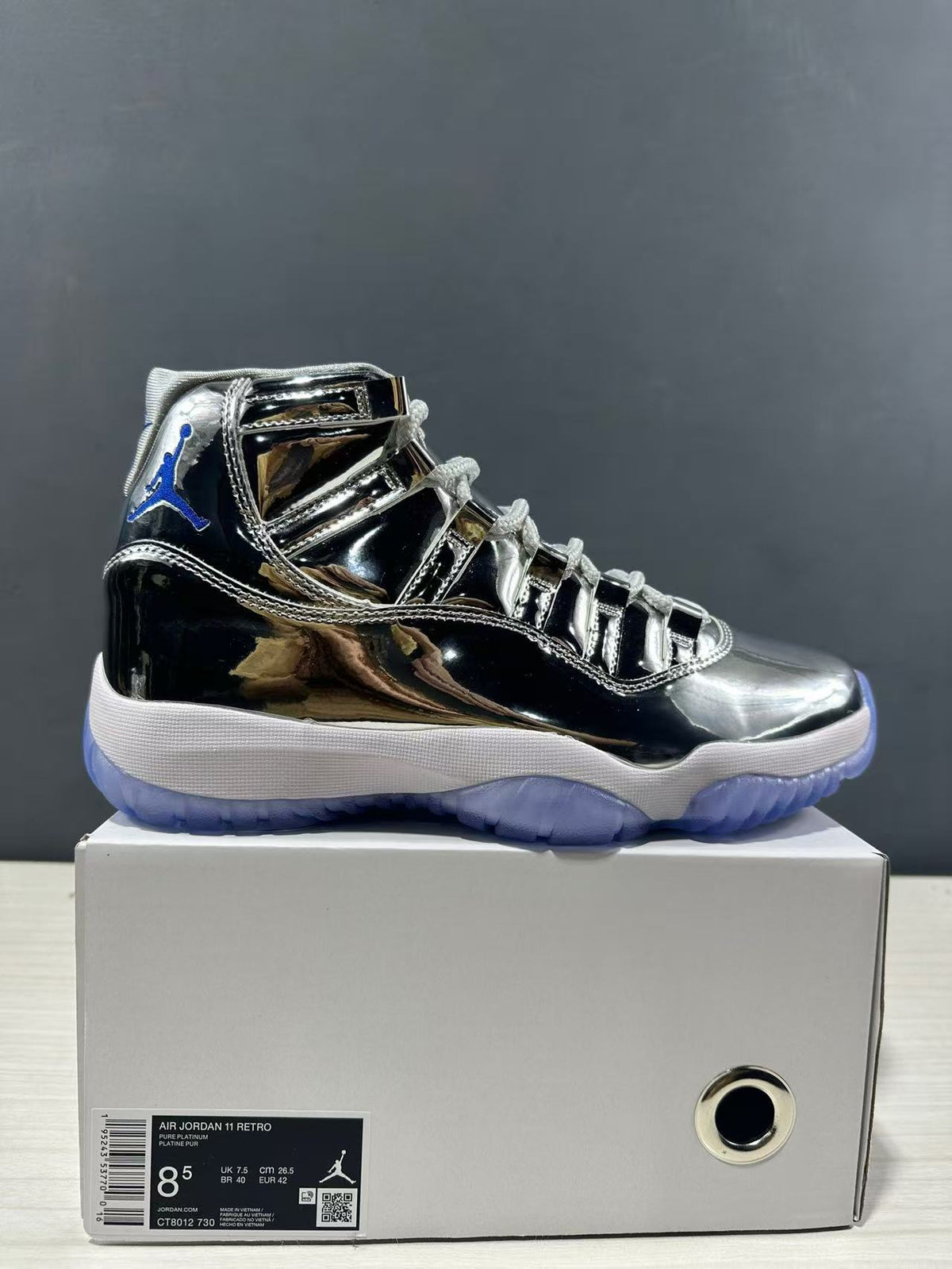 Side view of a reflective silver and black Air Jordan 11 sneaker on its box; the box label shows size 8.5, EU 42, CM 26.5, and style code CT8012 730.