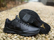 Black sneakers shown from above and below, highlighting the patterned rubber outsole and gold outsole logo.
