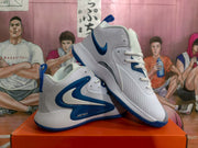 Angled view of white and blue Nike basketball sneakers showing the side branding, perforated panels, heel pull tabs, and textured outsole.