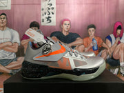 Side view of a silver basketball shoe with an orange Swoosh, midfoot strap, and speckled black midsole.
