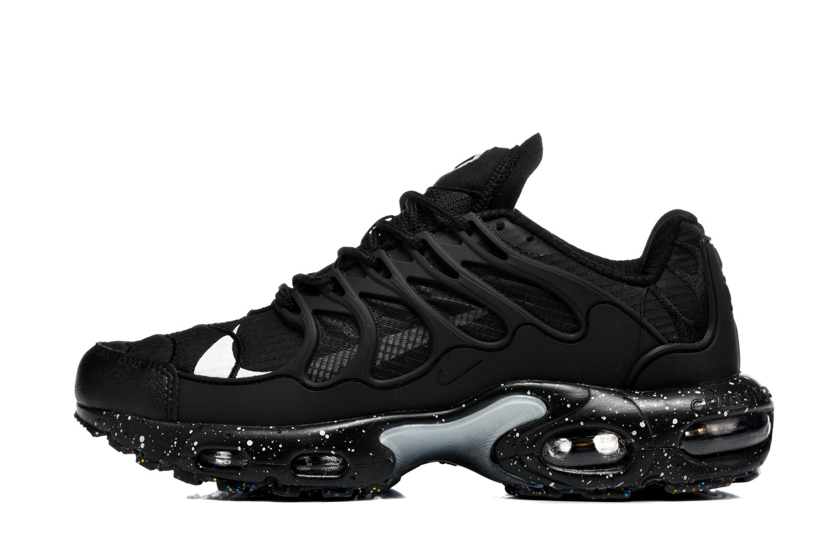 Side view of black Nike Air Max Plus sneakers with a layered mesh upper, sculpted overlay cage, visible cushioning, and a speckled midsole.