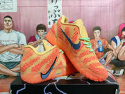 Pair of orange and tan Nike basketball sneakers displayed on a shoebox, showing the side profile, blue Swoosh branding, mint-green laces, and translucent orange outsole.