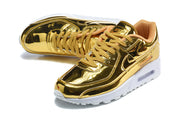 Top and angled views of gold-tone Nike Air Max 90 QS sneakers with tan laces.