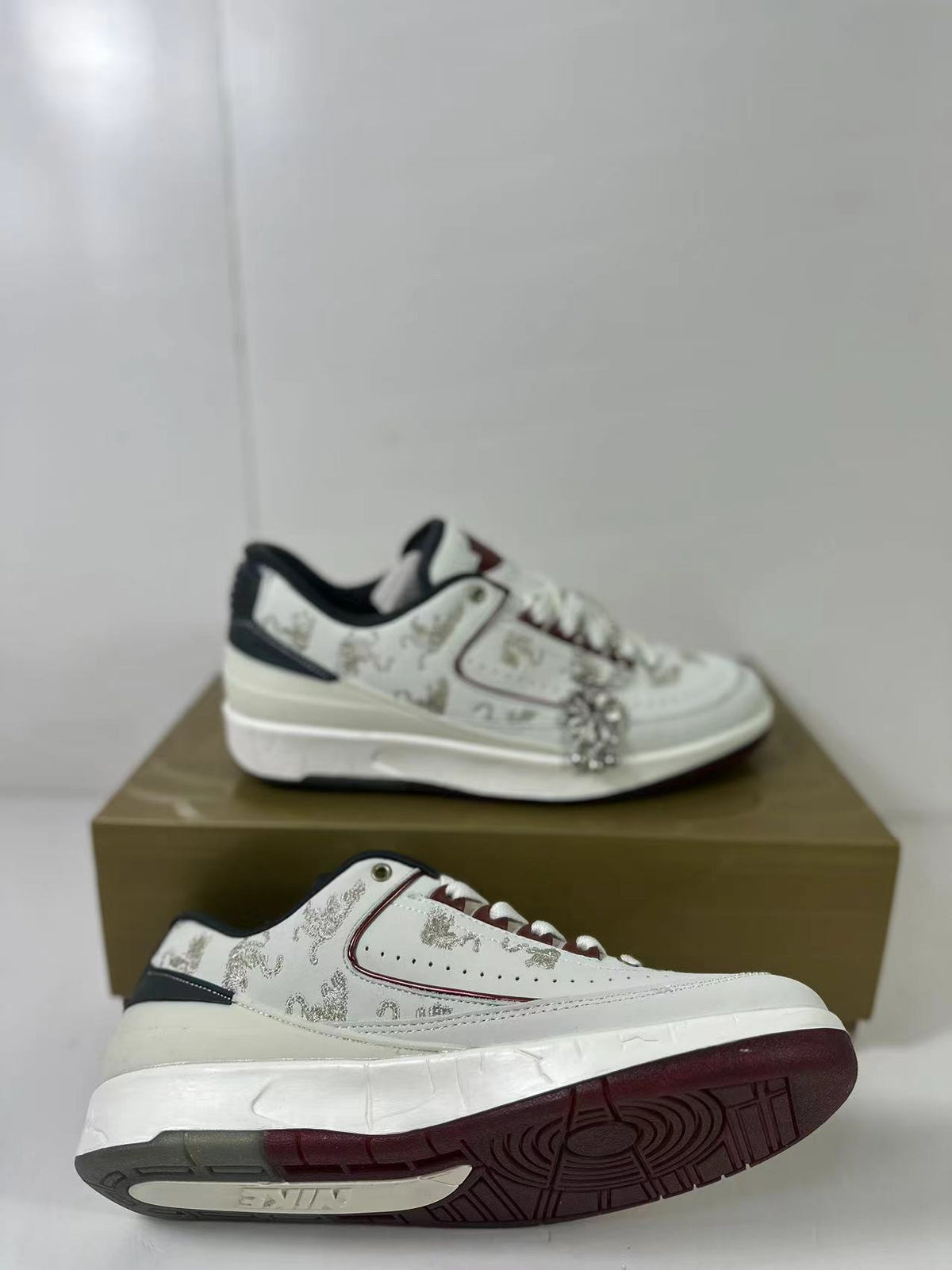 Side view of white Jordan low-top sneakers with dragon-pattern detailing, dark accents, and a decorative hanging charm on a box.