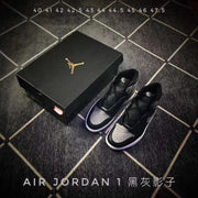 Pair of black and grey high-top sneakers beside a shoebox, with overlaid size text and Air Jordan 1 Shadow wording.