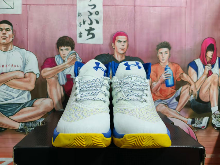 Front view of white high-top basketball sneakers with blue Under Armour tongue logos, patterned laces, and yellow outsole sections.
